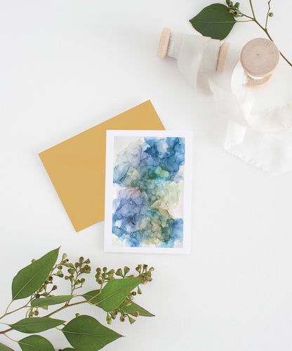 Greeting card with colorful envelope laying on a white surface with green leaves and cotton ribbon wound on wooden spools.