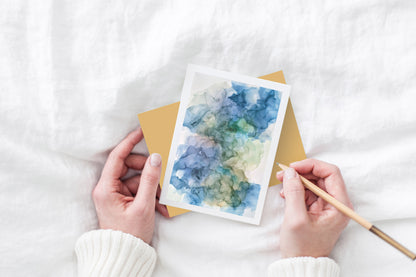 Greeting card and colorful envelope, seen from above, held by two hands along with a pencil, ready to write, against a white comforter.