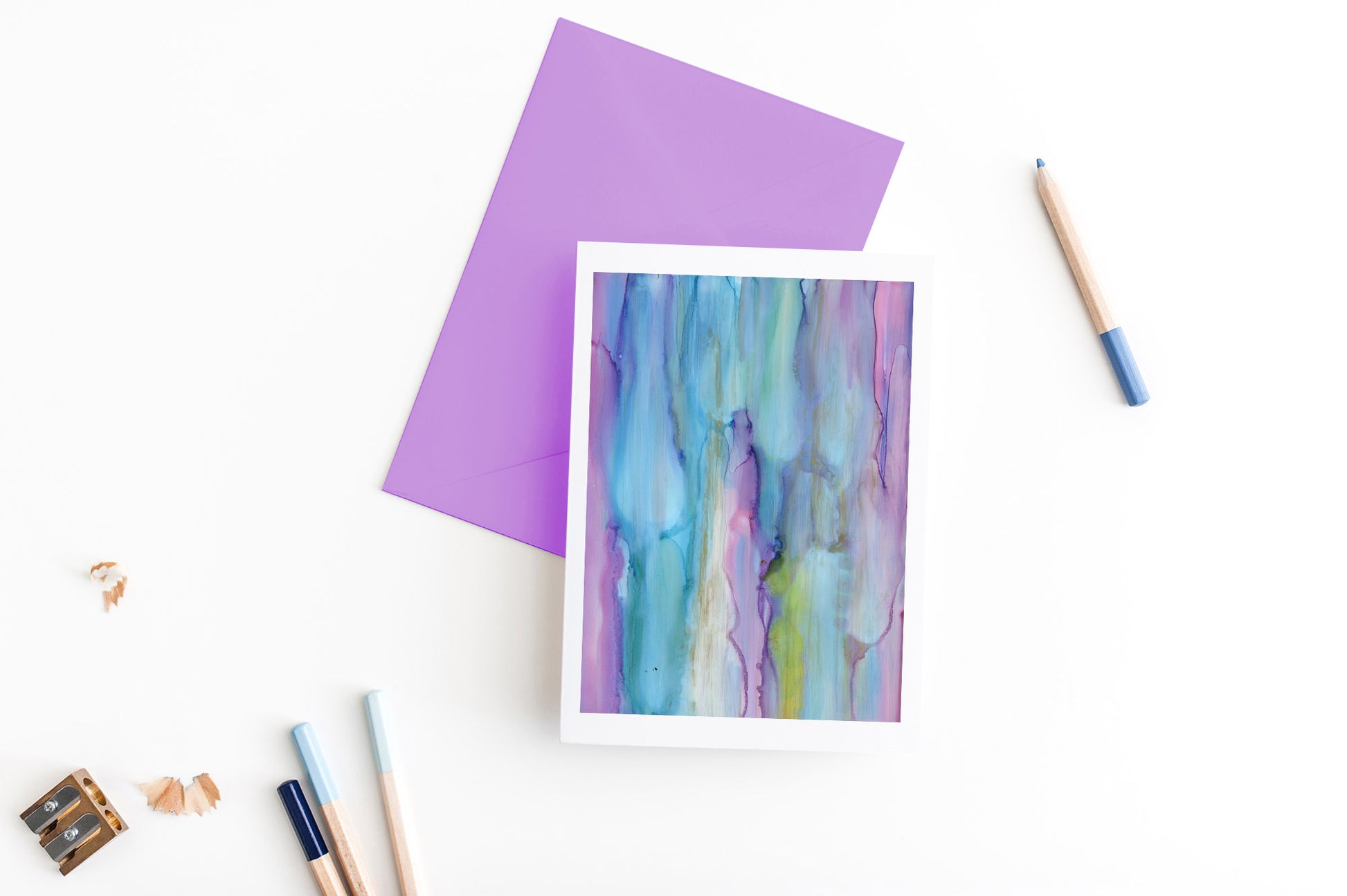 Greeting card, seen from above, with a colored, square flap envelope, pencils, pencil sharpener and shavings on a white background.