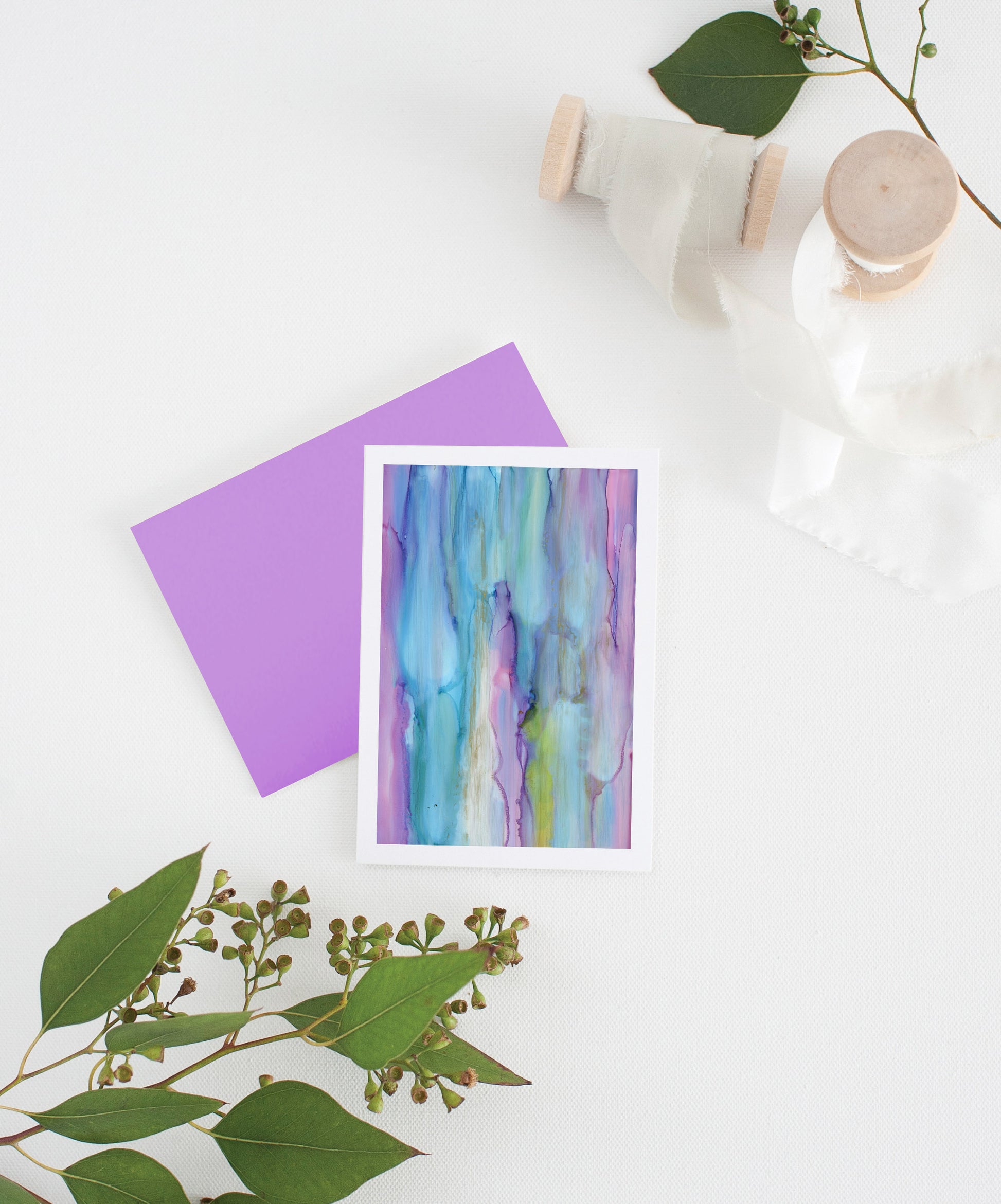 Greeting card with colorful envelope laying on a white surface with green leaves and cotton ribbon wound on wooden spools.