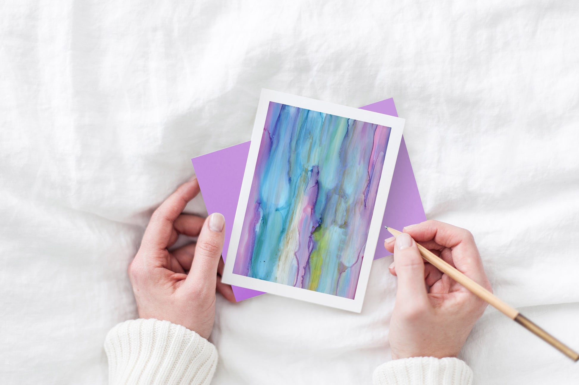 Greeting card and colorful envelope, seen from above, held by two hands along with a pencil, ready to write, against a white comforter.