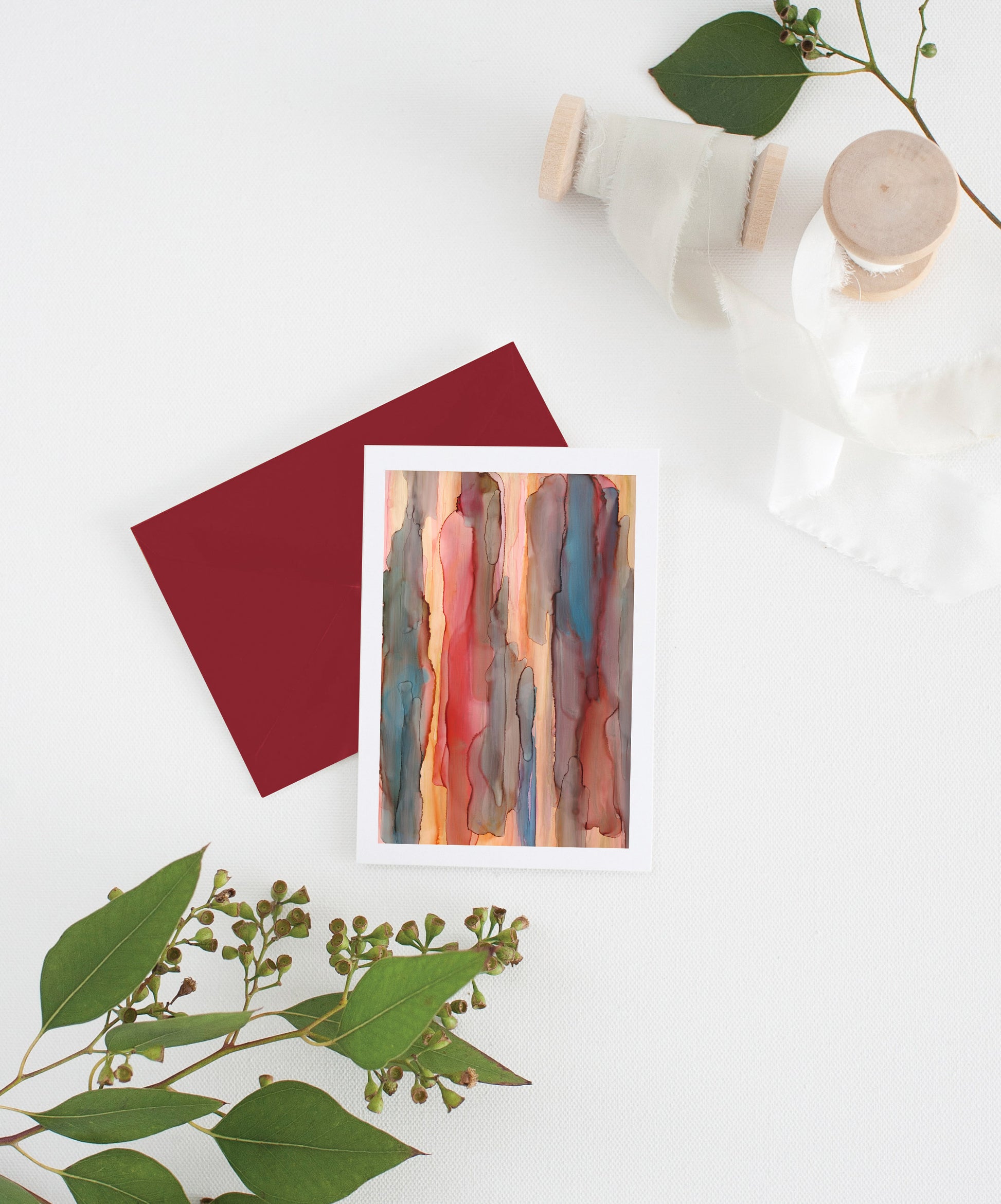 Greeting card with colorful envelope laying on a white surface with green leaves and cotton ribbon wound on wooden spools.