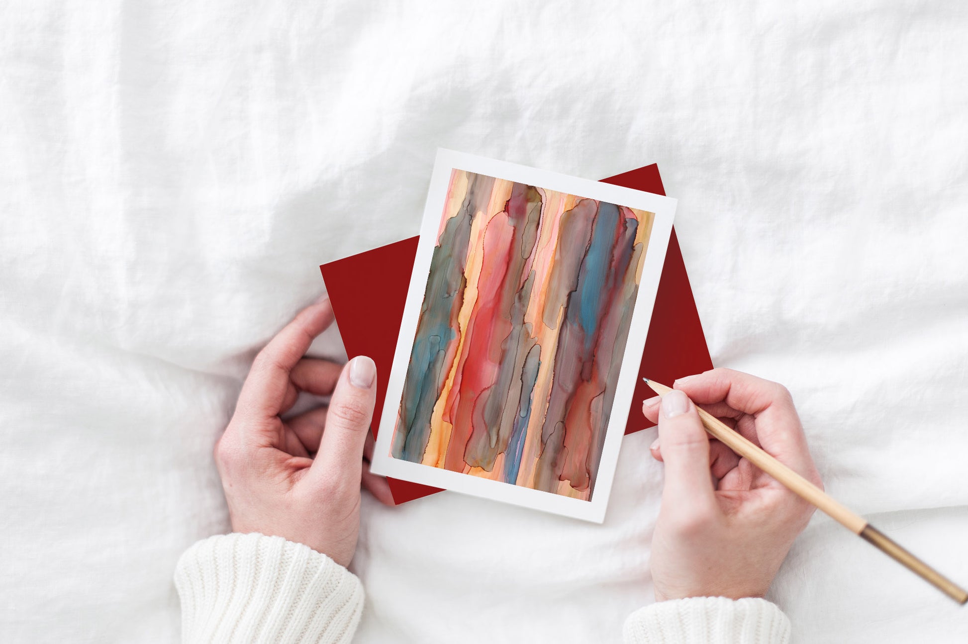 Greeting card and colorful envelope, seen from above, held by two hands along with a pencil, ready to write, against a white comforter.