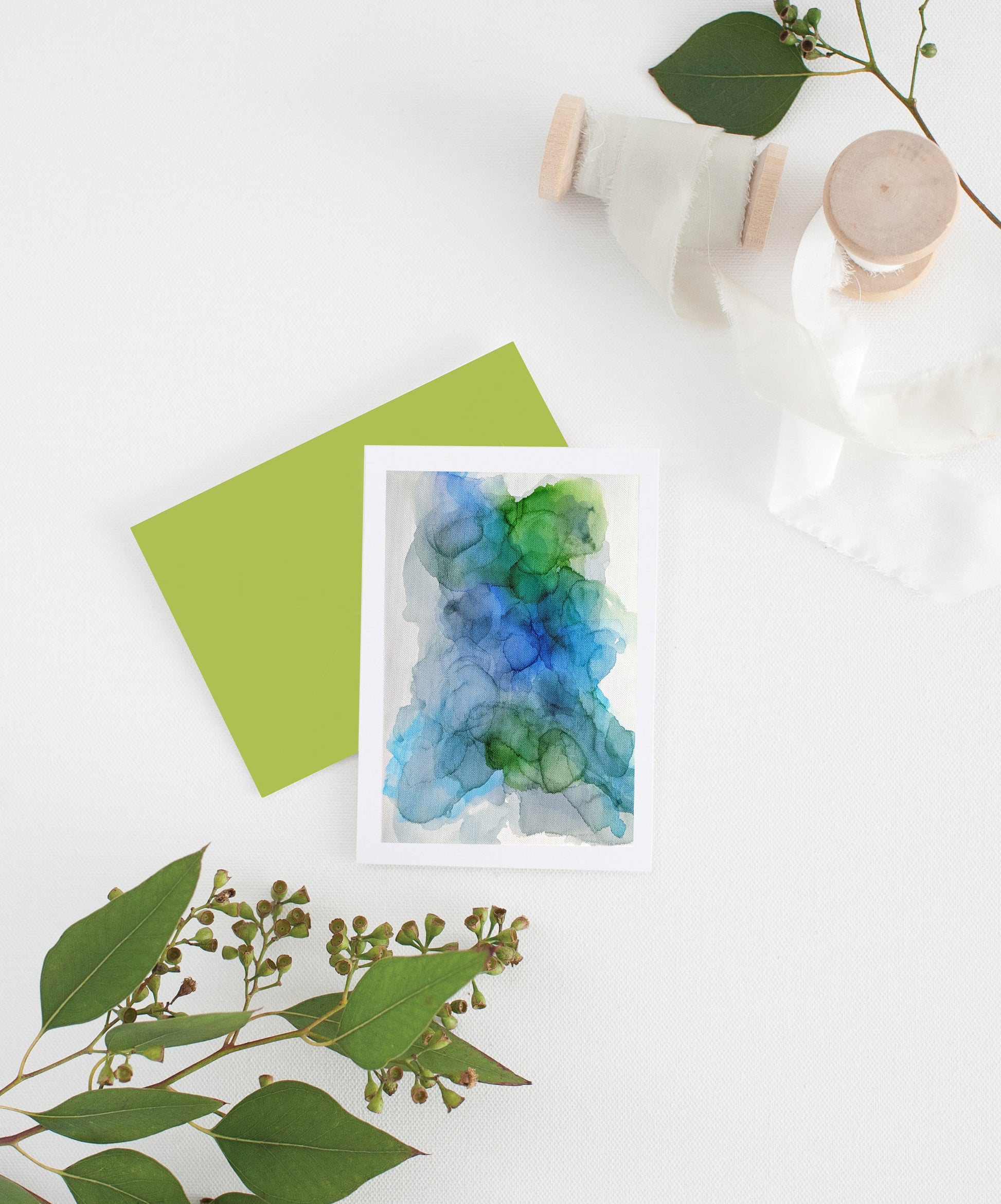 Greeting card with colorful envelope laying on a white surface with green leaves and cotton ribbon wound on wooden spools.