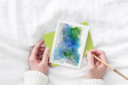 Greeting card and colorful envelope, seen from above, held by two hands along with a pencil, ready to write, against a white comforter.