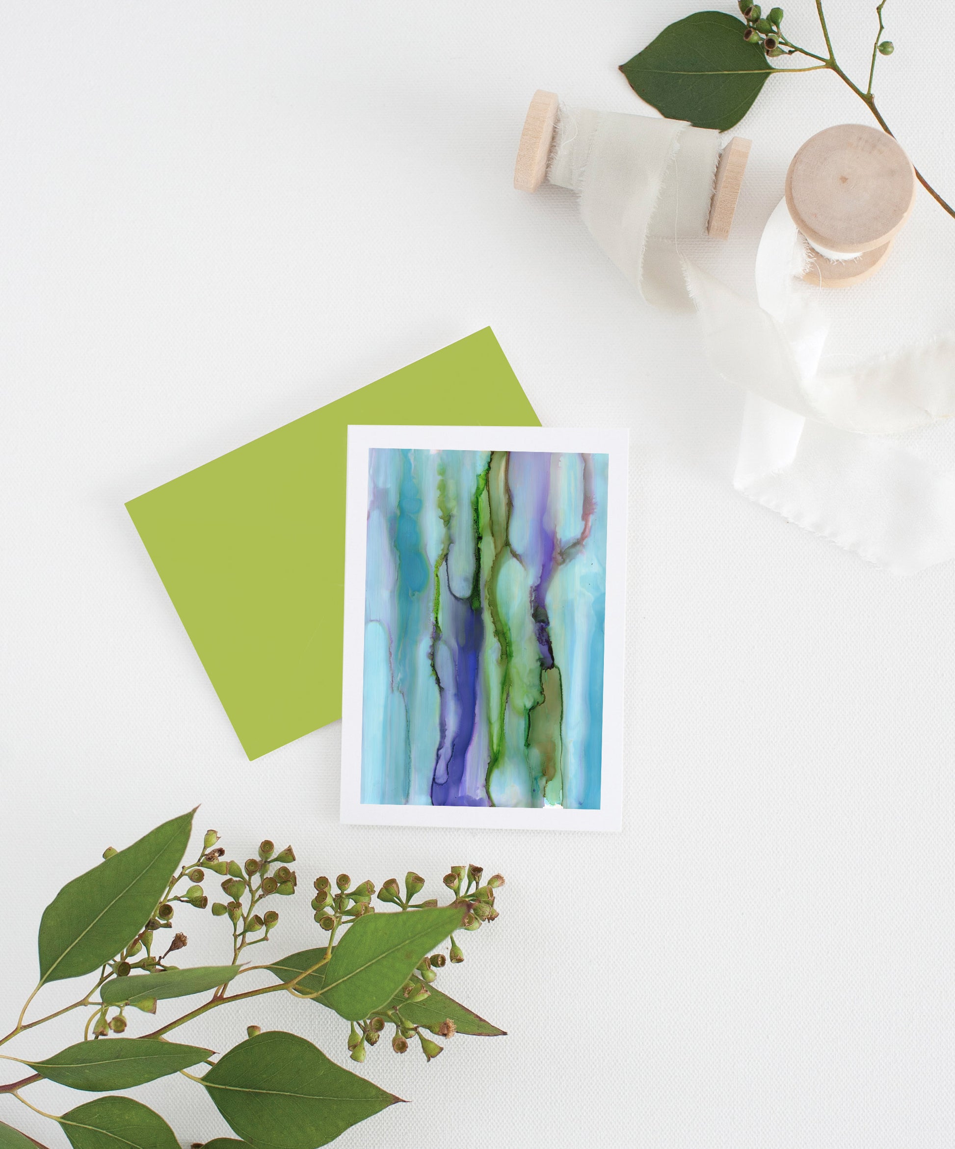 Greeting card with colorful envelope laying on a white surface with green leaves and cotton ribbon wound on wooden spools.