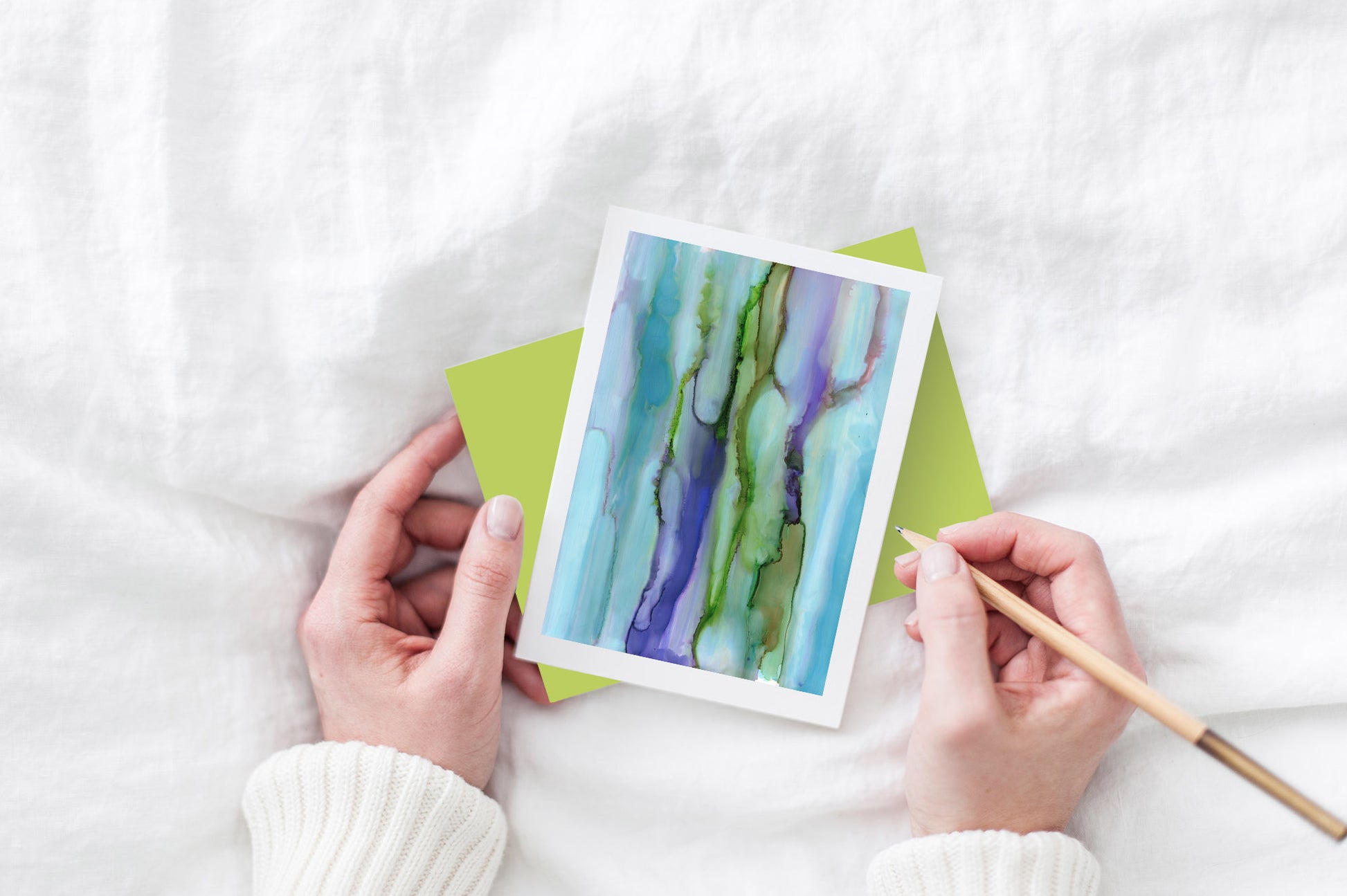 Greeting card and colorful envelope, seen from above, held by two hands along with a pencil, ready to write, against a white comforter.