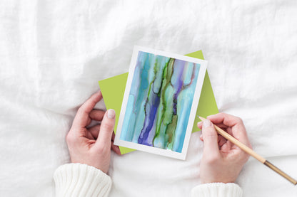 Greeting card and colorful envelope, seen from above, held by two hands along with a pencil, ready to write, against a white comforter.