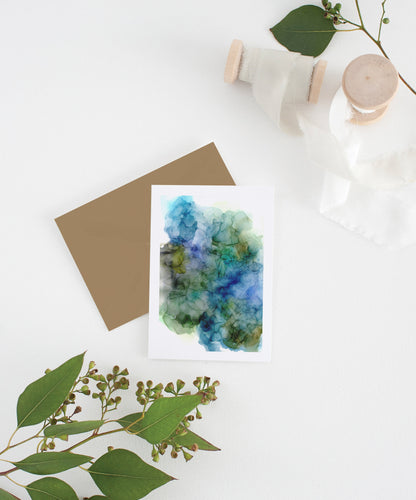 Greeting card with colorful envelope laying on a white surface with green leaves and cotton ribbon wound on wooden spools.