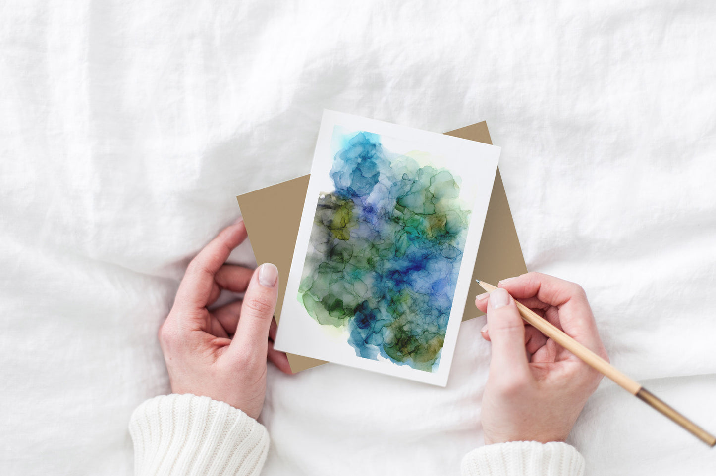 Greeting card and colorful envelope, seen from above, held by two hands along with a pencil, ready to write, against a white comforter.