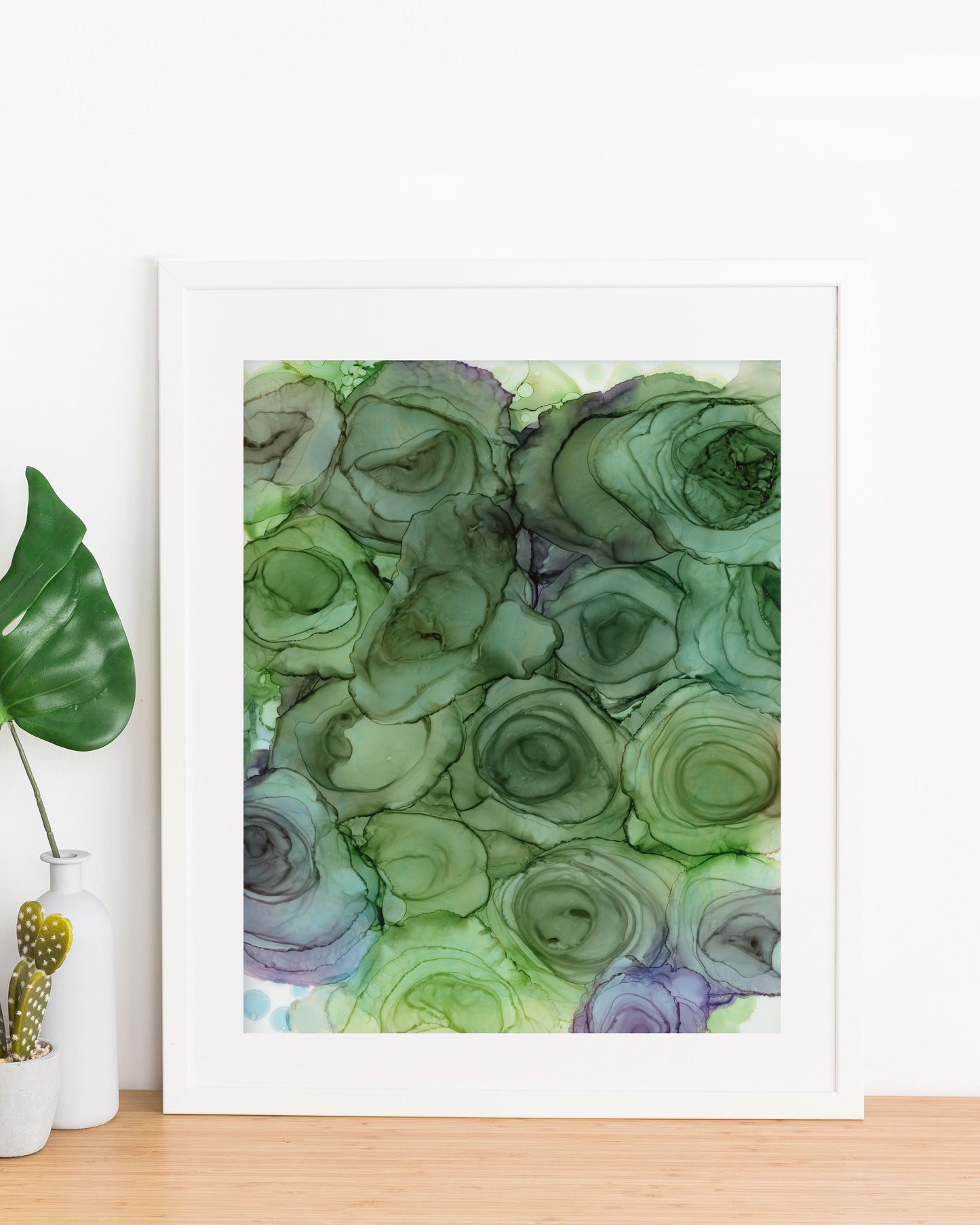 Verdant Blooms painting displayed in a white frame propped up against a white wall, on a wood table with a small vase and planter on the left.