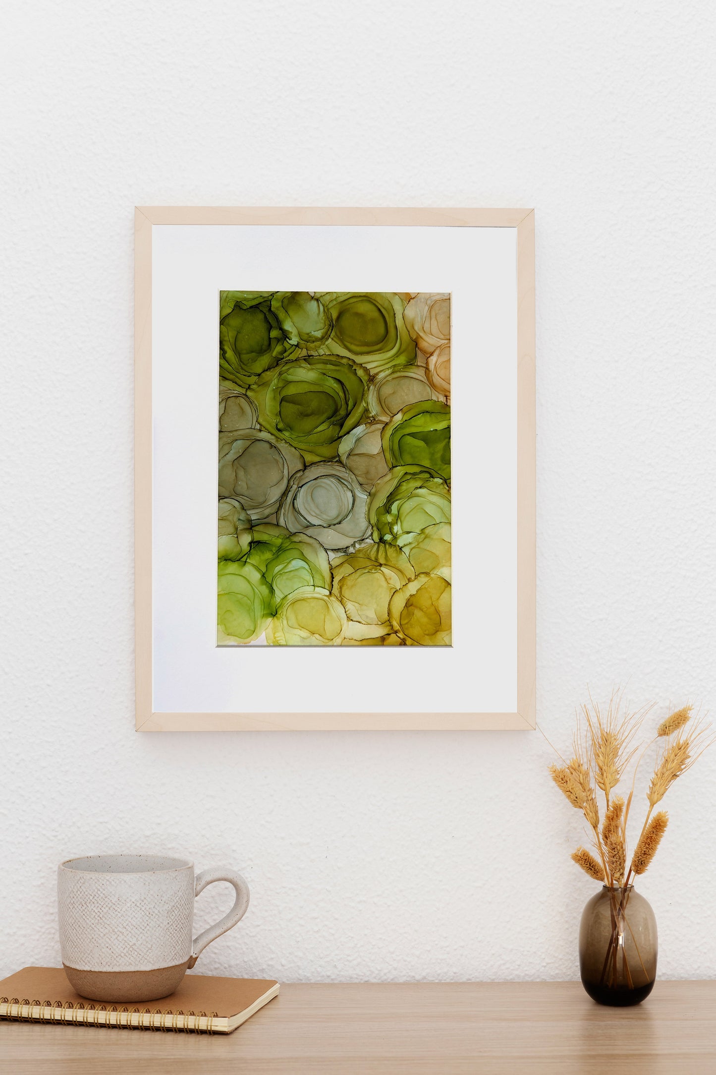 Digital mockup of Viridescent Blooms framed in light wood  hanging on a slightly textured white wall.  The table top below holds a small vase with grains, a ceramic mug and a wirebound notebook.