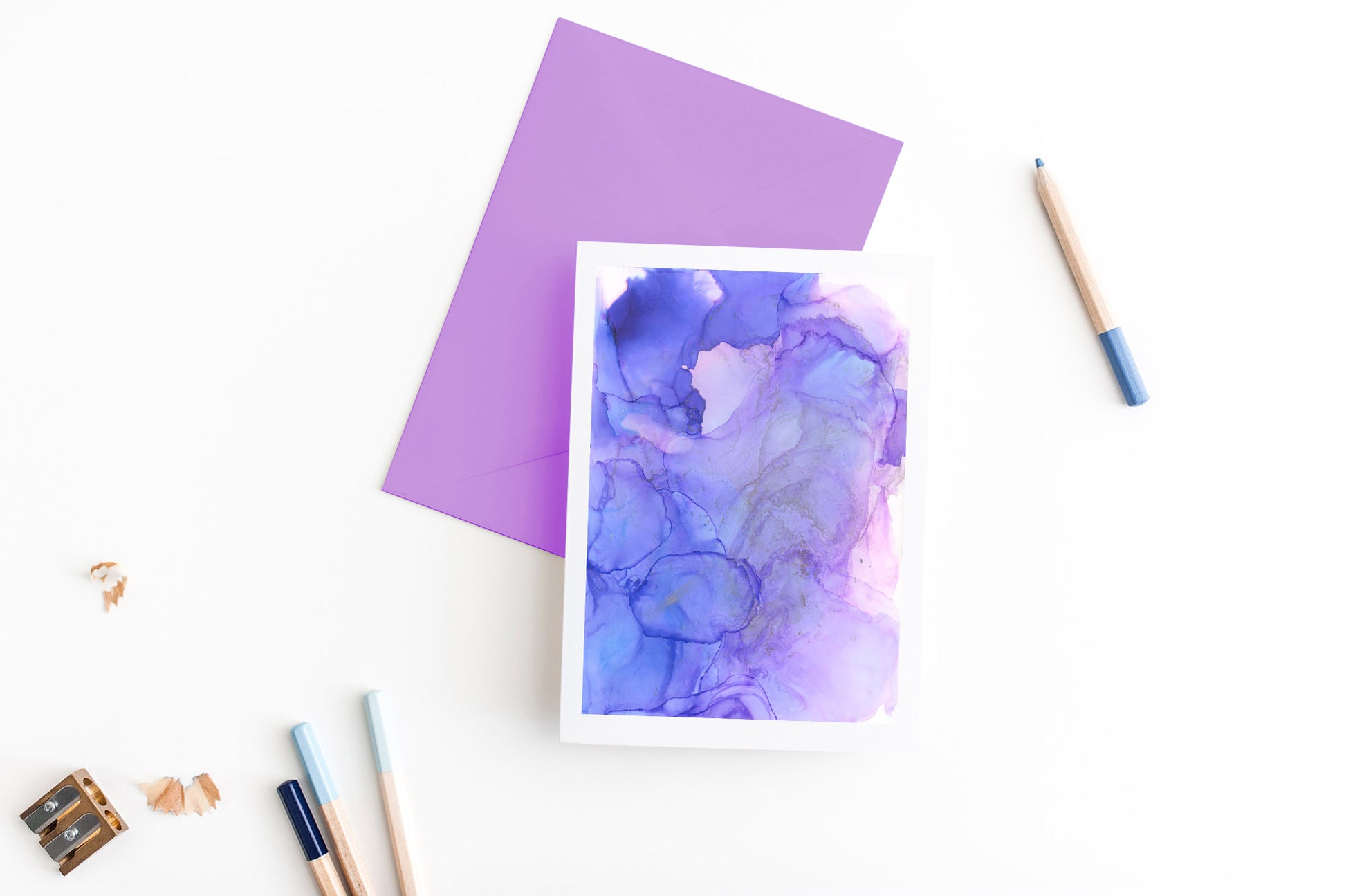 Greeting card, seen from above, with a colored, square flap envelope, pencils, pencil sharpener and shavings on a white background.