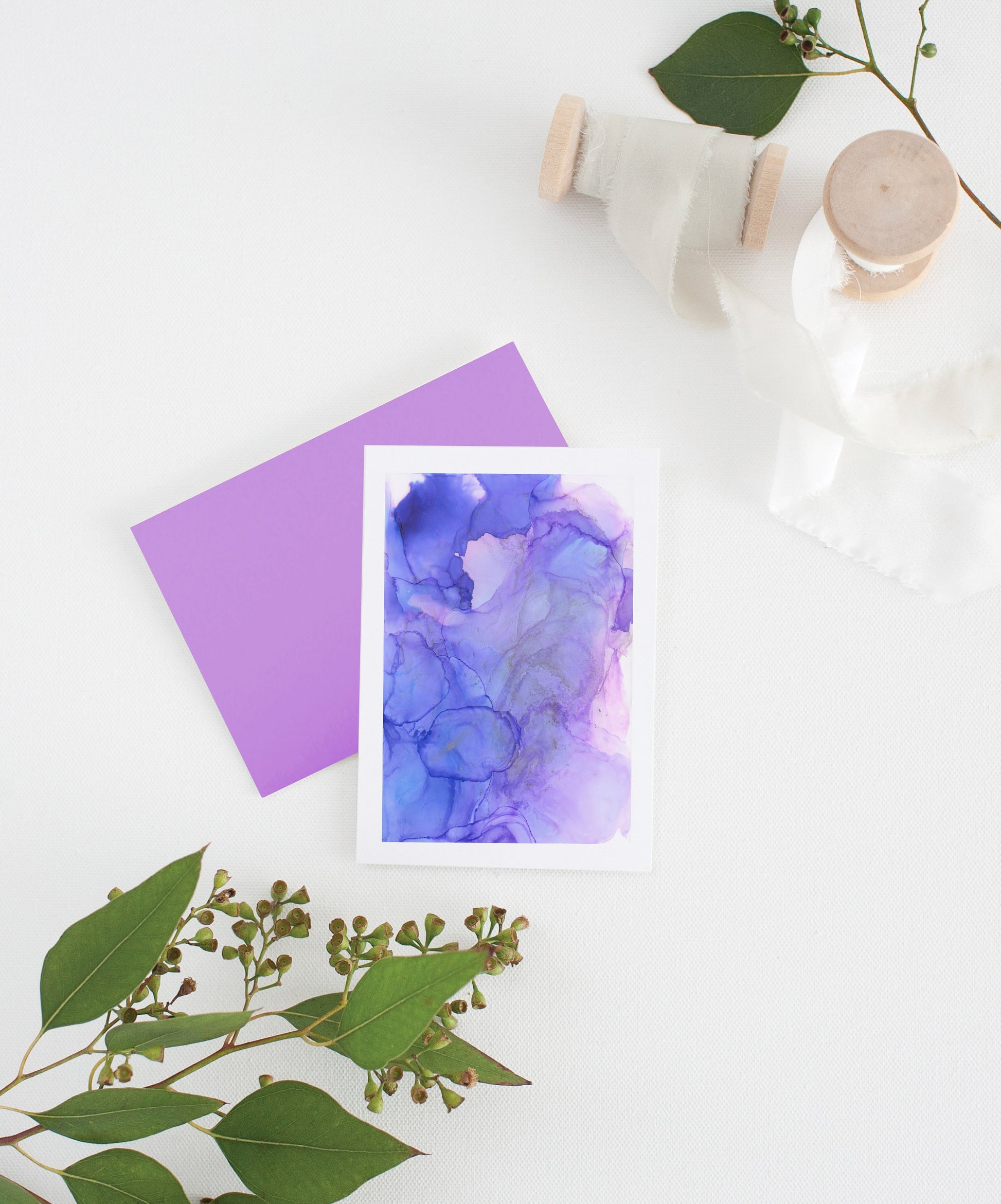 Greeting card with colorful envelope laying on a white surface with green leaves and cotton ribbon wound on wooden spools.