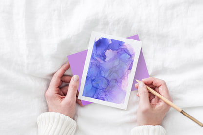 Greeting card and colorful envelope, seen from above, held by two hands along with a pencil, ready to write, against a white comforter.