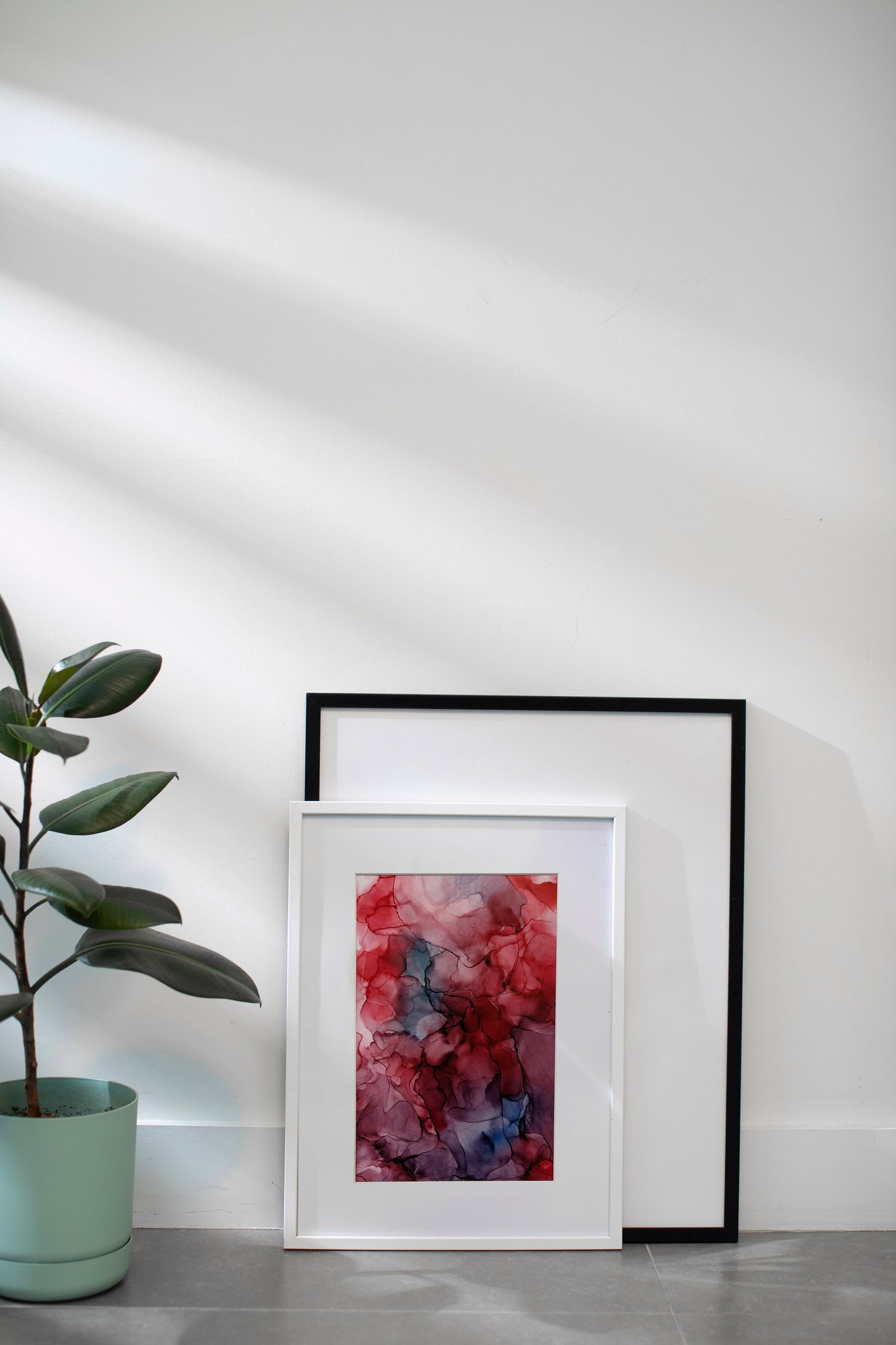 Zoomed out view digital mockup of Wonderful and Unexpected Ways framed in white leaning on grey tiles up against a white wall and another empty black frame. A plant in a mint green plant sits off to the side.