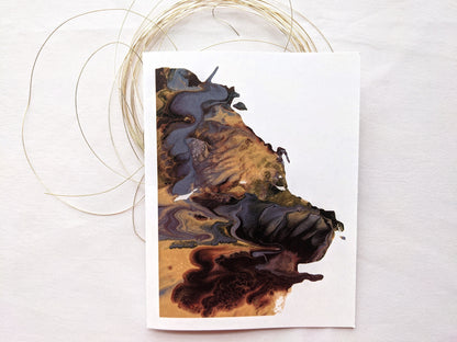 Greeting card showing an abstract painting in golds, dusty blues, and browns created from squishing acrylic paint between pieces of cardboard. It has thin gold wire looped beneath it and sits on a white cloth.