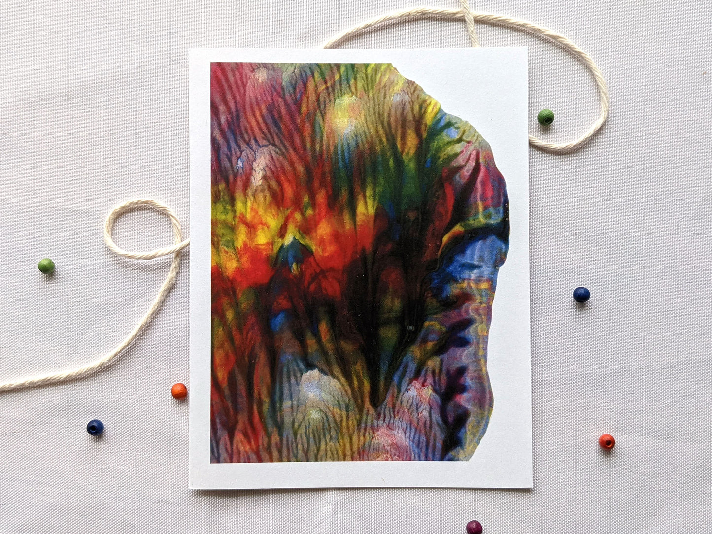 A greeting card with rainbow colored abstract art sits on a white cloth background with cooking twine and small colorful wooden beads.