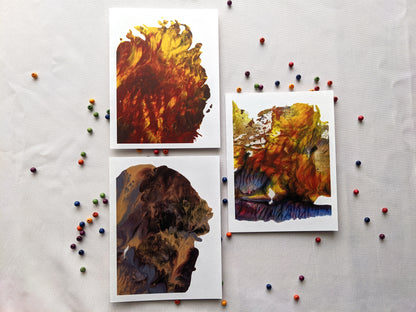 Three greeting cards with abstract paintings laying on a white cloth background surrounded by small colorful wooden beads.