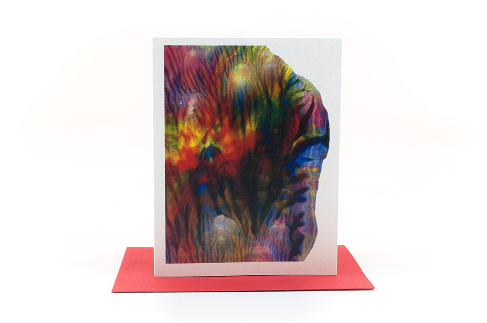 Greeting card showing an abstract painting in rainbow colors created from squishing acrylic paint between pieces of cardboard. It sits on a red envelope.