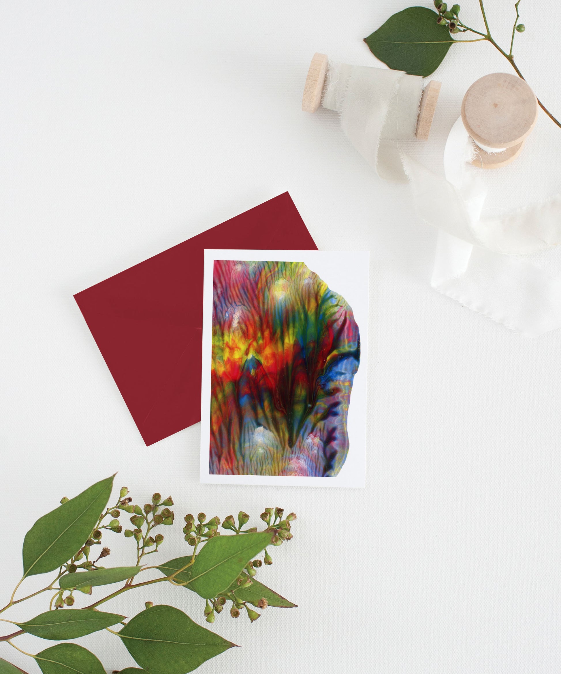 An abstract painting greeting card sits with a red envelope on a white background.  There are leaves and two small spools of white fabric ribbon near it.