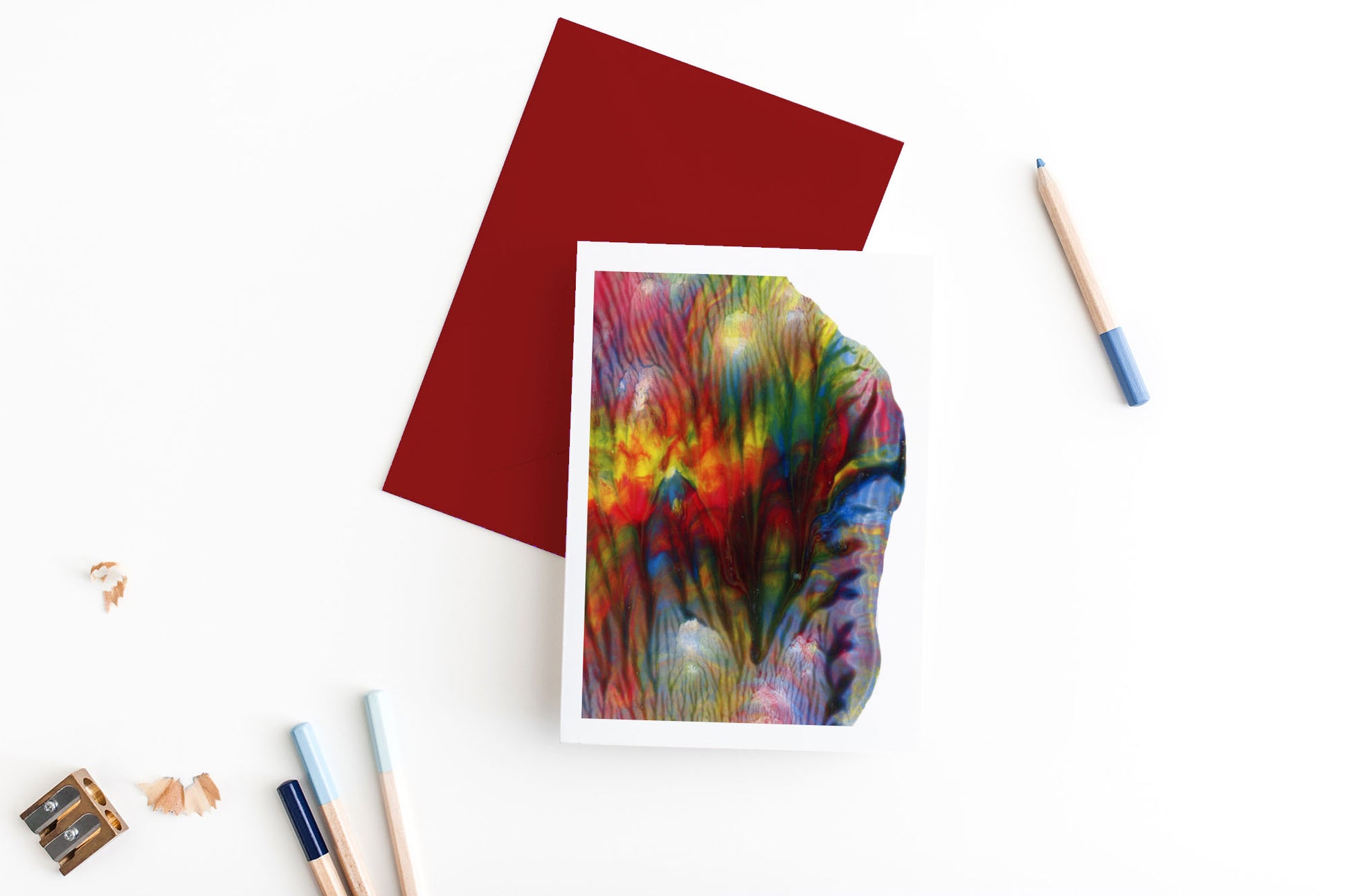 Greeting card showing an abstract painting in rainbow colors created from squishing acrylic paint between pieces of cardboard. It sits on a red envelope with 4 colored pencils, a sharpener and shavings on a white background.