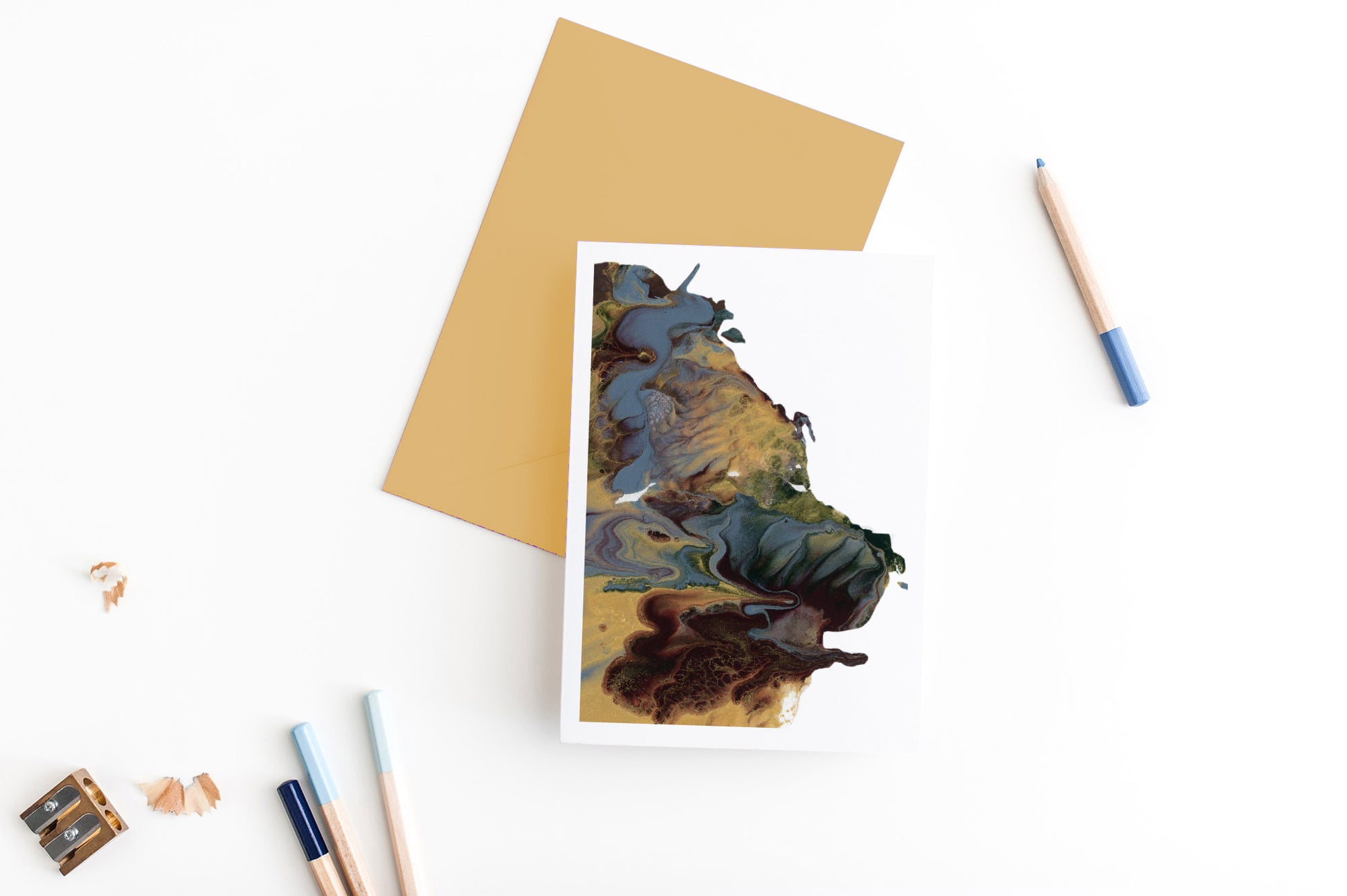 Greeting card showing an abstract painting in golds, dusty blues, and browns created from squishing acrylic paint between pieces of cardboard. It sits on a gold envelope with 4 colored pencils, a sharpener and shavings on a white background.