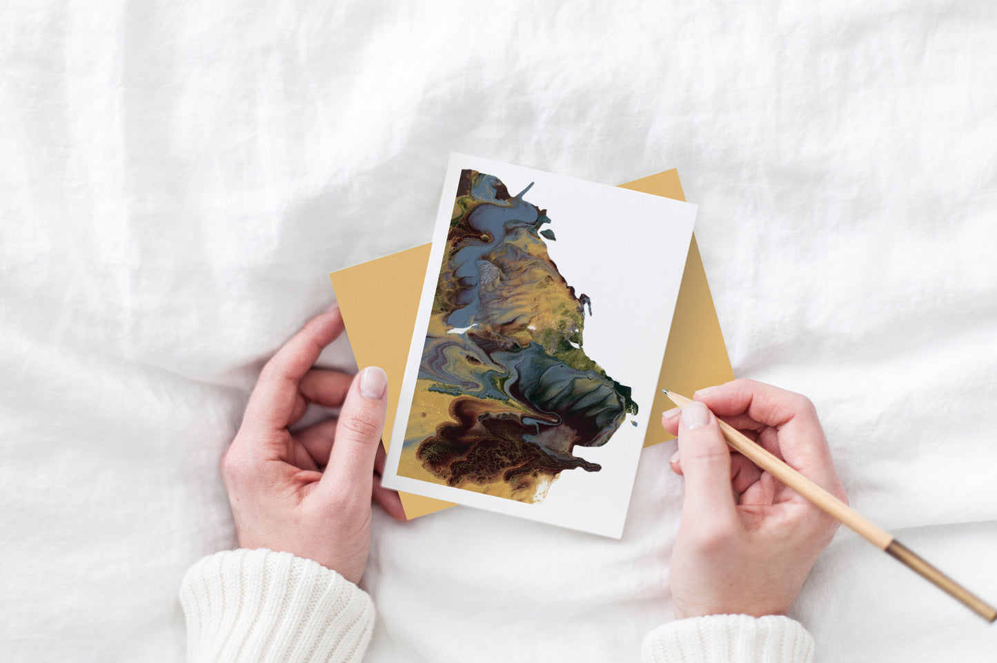 The abstract painting greeting card sits with a gold envelope on white bedding with a women's hands on either side of it. She's holding a pencil and you can see her white sweater sleeves.
