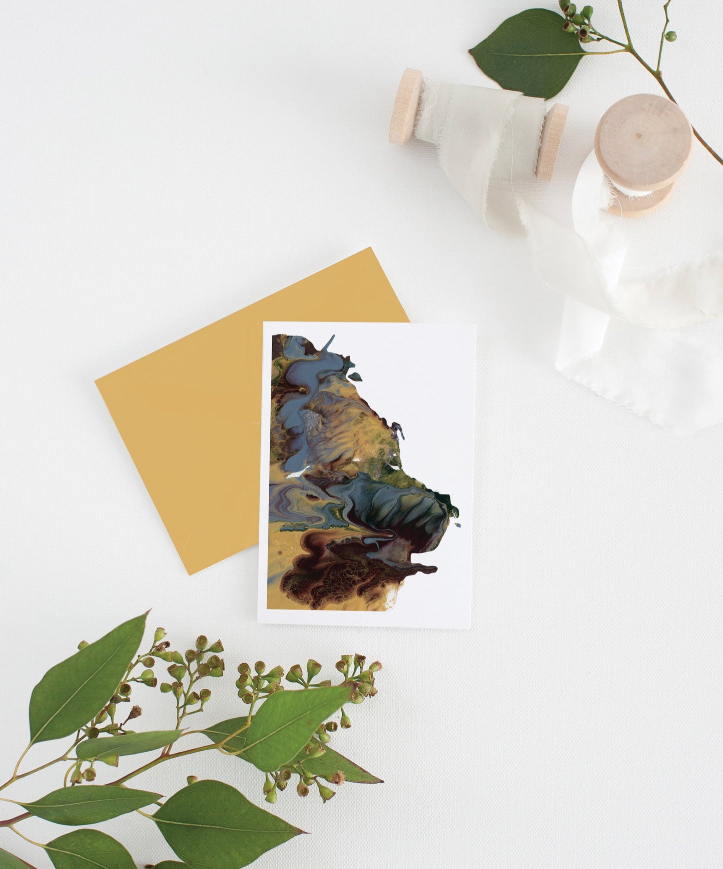 An abstract painting greeting card sits with a gold envelope on a white background.  There are leaves and two small spools of white fabric ribbon near it.
