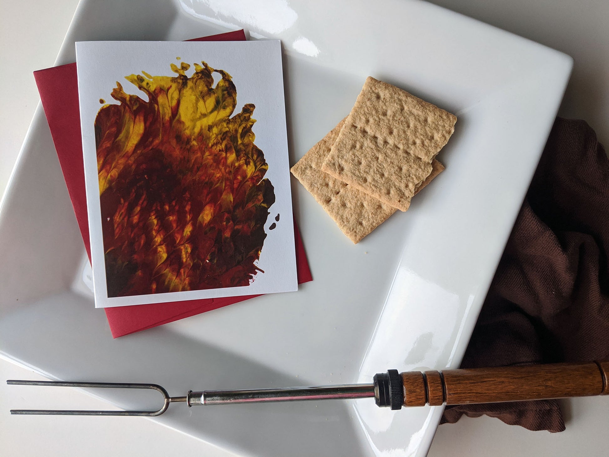 Greeting card with abstract art that looks like flames sitting on a square, white plate, with a red envelope, graham crackers, a smore roasting stick and a brown cloth napkin.