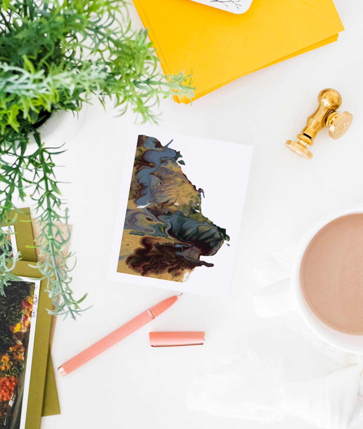 Greeting card showing an abstract painting in golds, dusty blues, and browns created from squishing acrylic paint between pieces of cardboard. It sits on a white background amongst a green plant,  a stack of yellow envelopes, a pink pen, a gold wax seal, and a white mug filled with coffee.