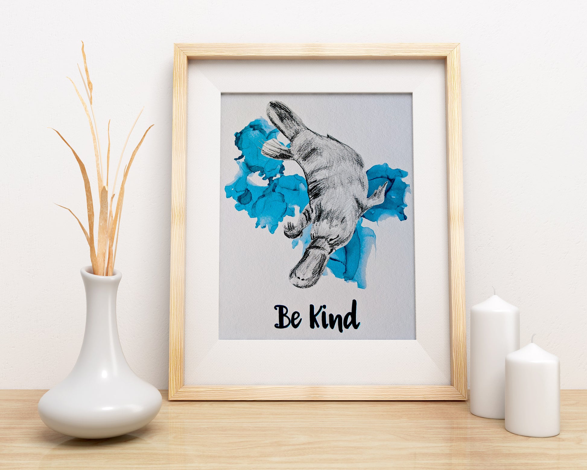 Digital mockup of an animal art print with foil accents framed in wood standing on a wooden surface leaning against a white wall.  Two pillar candles and a vase with dried stems sit to the sides.