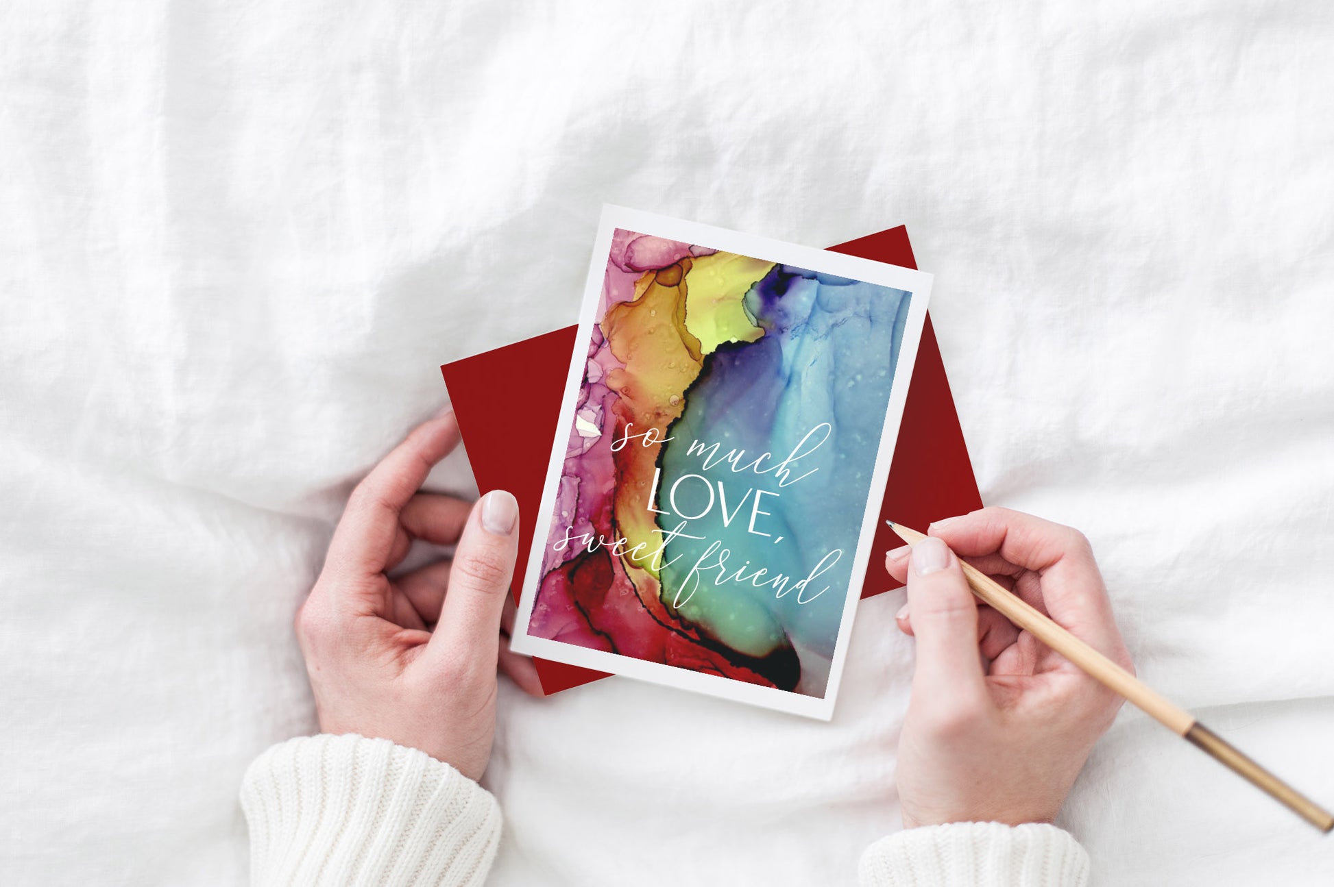 Greeting card and colorful envelope, seen from above, held by two hands along with a pencil, ready to write, against a white comforter.